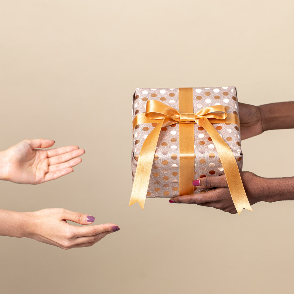 Dark-skinned hands present a gold polka dot gift with a bow to light-skinned hands reaching out to receive it.
